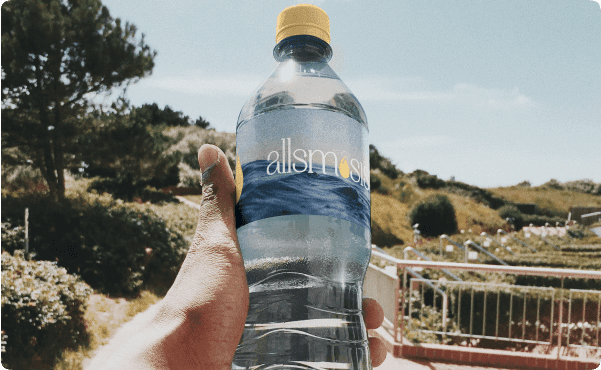 A hand holds a bottle of "allsmosis" water outdoors on a sunny day, with trees, grass, and a clear sky in the background, conveying a refreshing vibe.